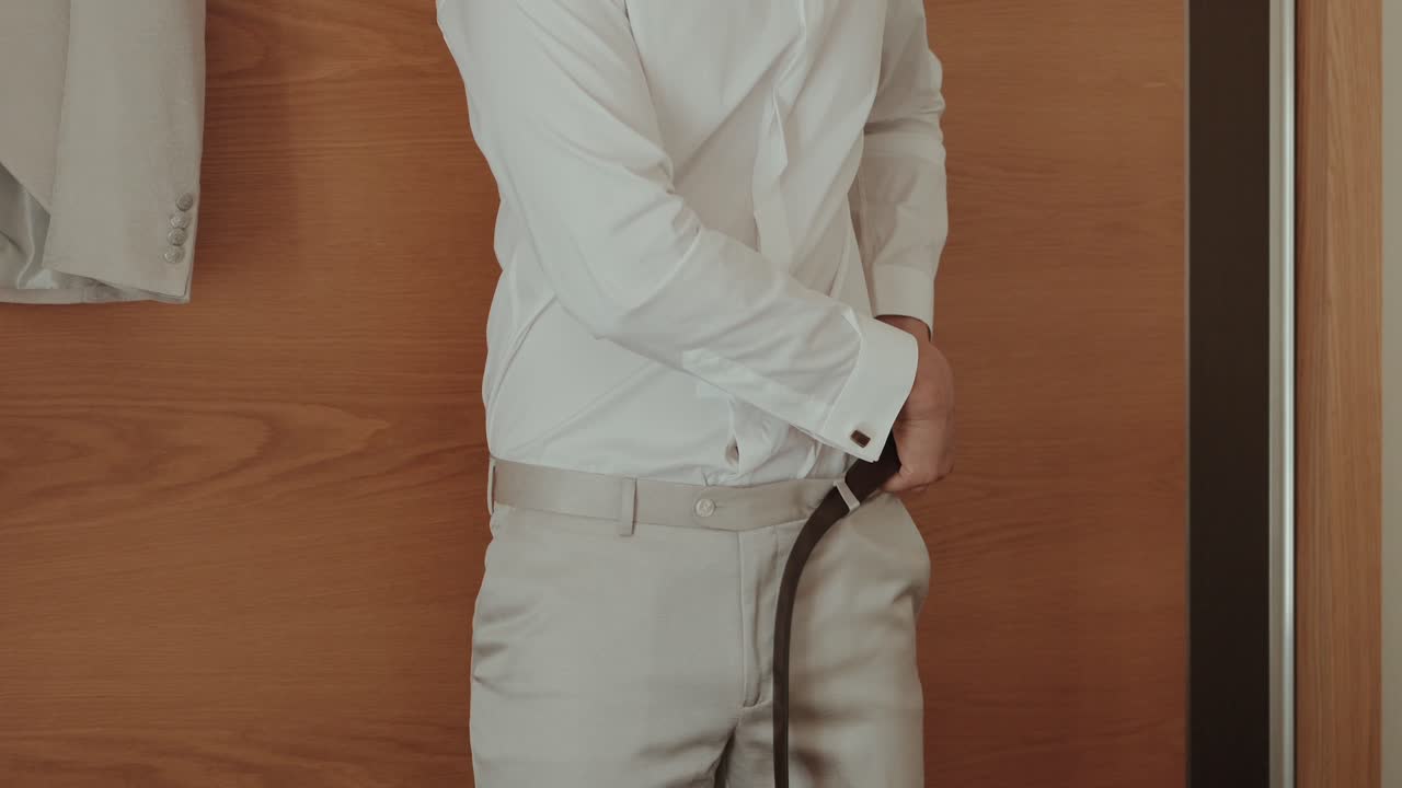 Close up of a man in a white shirt adjusting a brown belt on beige trousers indoors