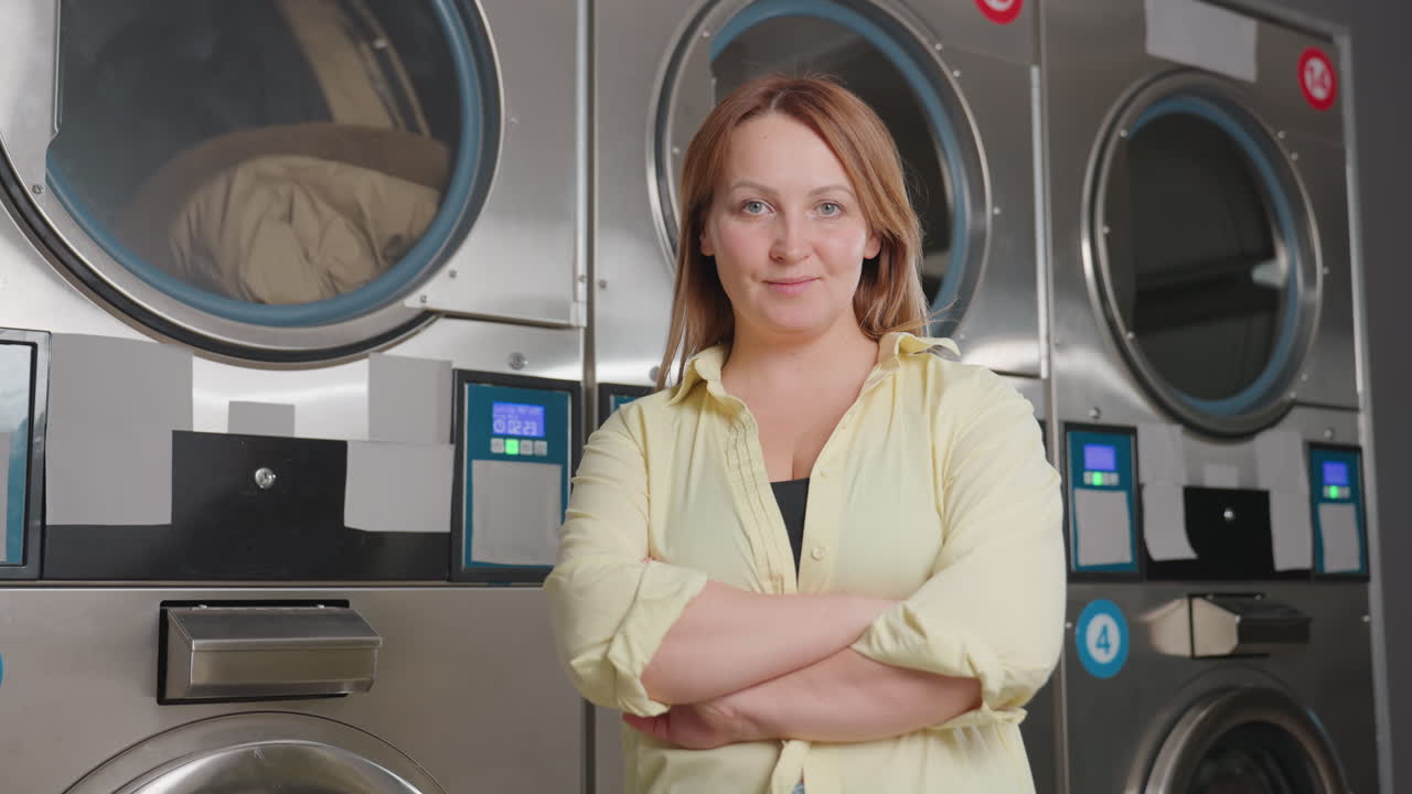 Laundry owner poses with hands folded, while standing before industrial cloth dryers, clothes rolling behind glass, stainless machines with glowing controls, modern laundromat showcasing service