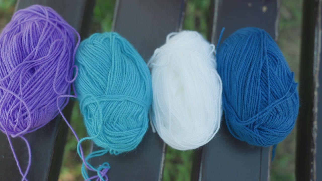 Primo piano di una mano che sistema gomitoli di lana colorati su una panchina all'aperto. La scena mostra gomitoli viola, blu, bianchi e di altri colori che vengono posizionati in sequenza su uno sfondo di fogliame verde. Morbido.