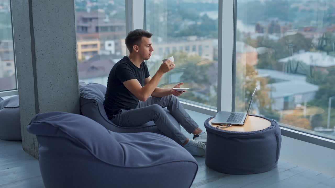 Young self-employed man having lunch break resting from work at laptop. Male drinking coffee and enjoying city view from a big window.