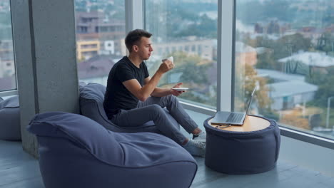young self-employed man having lunch break resting from work at laptop. male drinking coffee and enjoying city view from a big window.