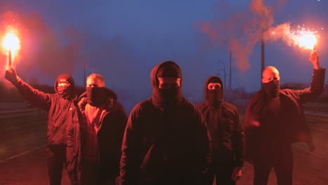 group of young men in balaclavas with red burning signal flare walking on the road under the bridge