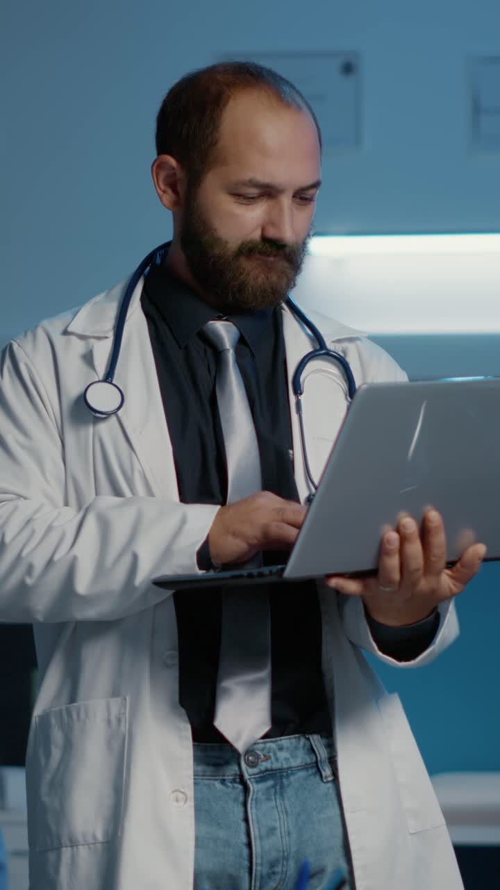 Vertical video: Portrait of smiling doctor wearing medical coat and stethoscope holding laptop computer