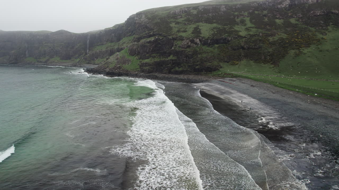 disparo aéreo de drones de olas en el océano atlántico rodando en una playa de grava con cascada en el fondo en la bahía de talisker en escocia