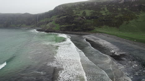 Toma-Aérea-De-Olas-Del-Océano-Atlántico-Rodando-En-Una-Playa-De-Grava-Con-Cascada-En-El-Fondo-En-La-Bahía-De-Talisker-En-Escocia