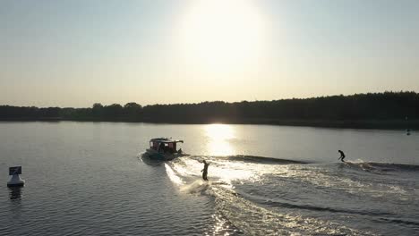 Two-surfers-on-surfboard-surfing-waves-behind-boat-in-sunset