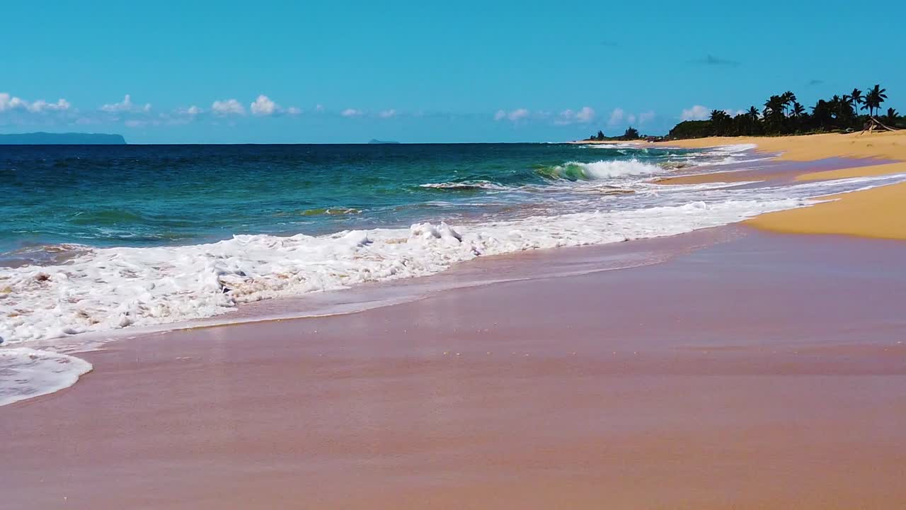 HD Hawaii Kauai, ripresa lenta in movimento a bassa quota con la spiaggia lungo il bordo inferiore verso la destra dell'inquadratura, con onde oceaniche che si infrangono e si estendono attraverso l'inquadratura da sinistra a destra, e un'isola in lontananza sulla sinistra dell'inquadratura