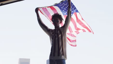 hombre afroamericano con gafas de sol sosteniendo una bandera estadounidense mientras está de pie cerca de la ventana