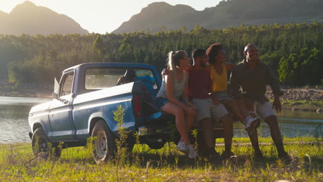 friends with backpacks sitting on tailgate of pick up truck on road trip by lake in countryside