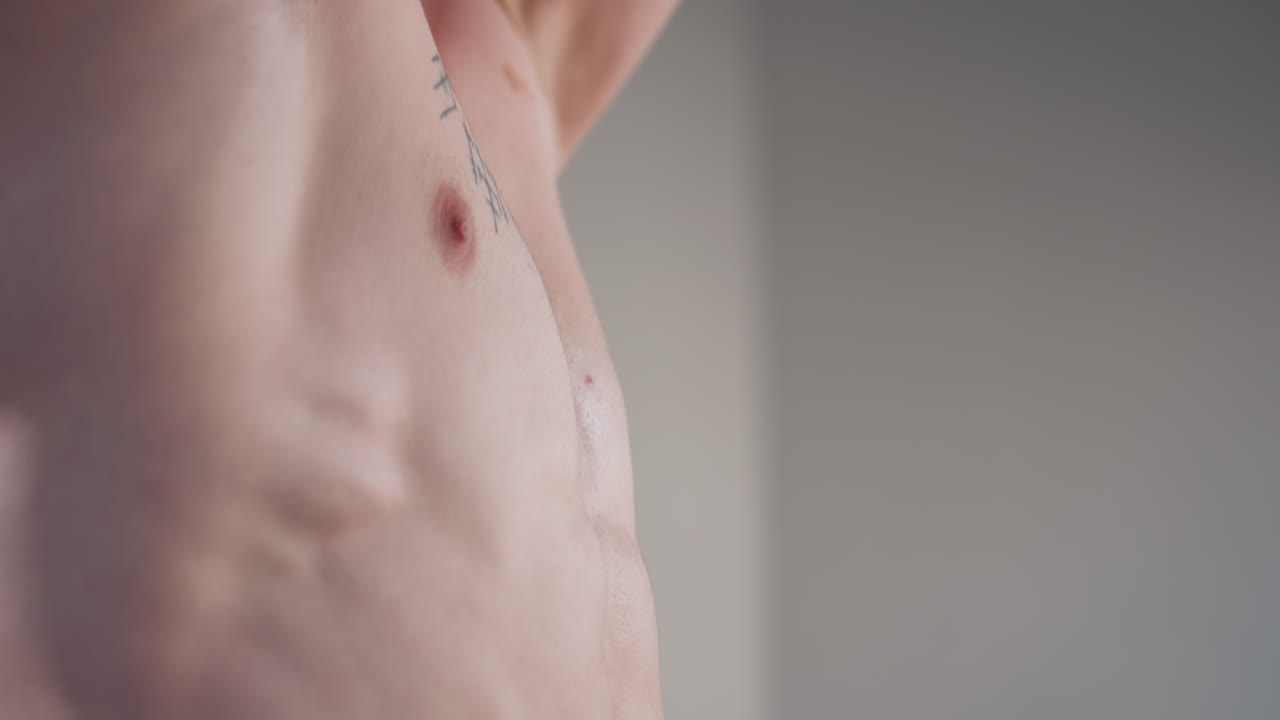 shirtless fair-skinned young man confidently shows defined upper body with arms raised behind head, revealing toned abs and chest tattoo while smiling and looking into camera in bright indoor setting