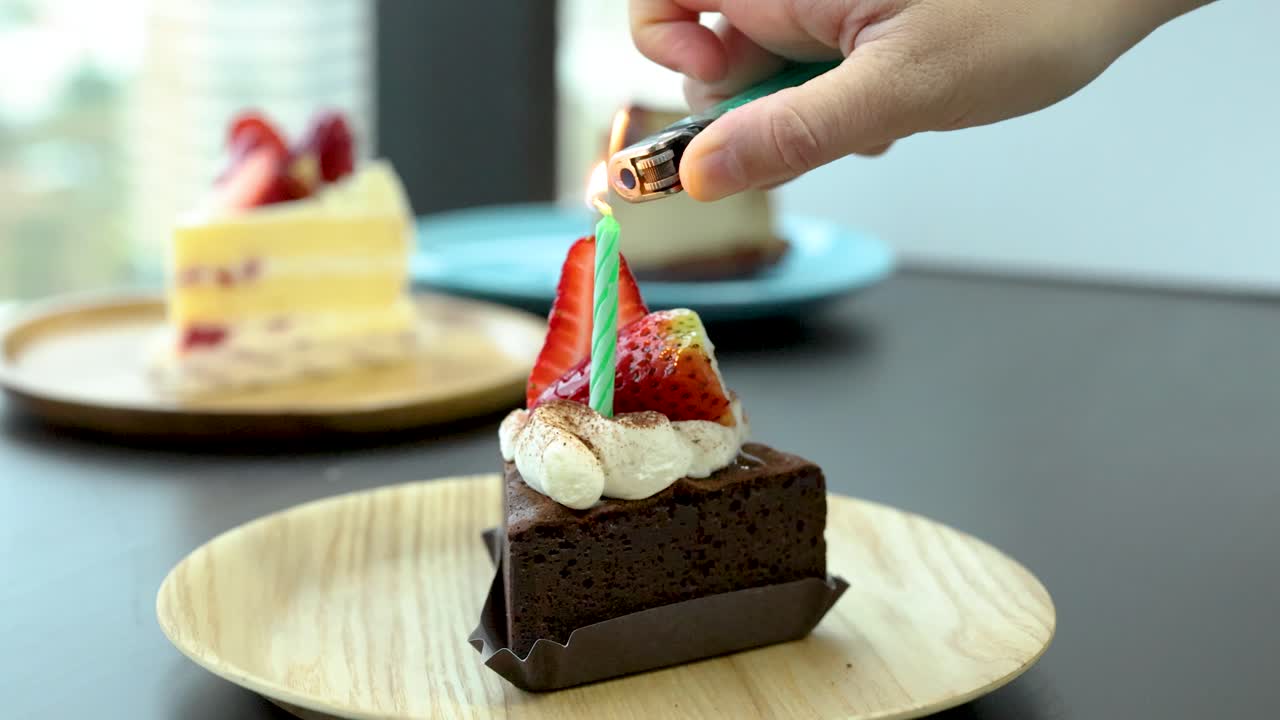 Hand lights candle on chocolate strawberry cake slice in bright, modern indoor setting, natural daylight