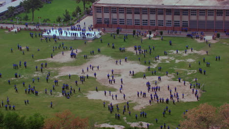 school students playing in the ground aerial zoom out of the ground, some running and having fun, students are in blue pants and shirts, school building in the background