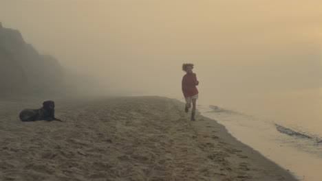 excited woman running at seashore in sunrise. happy girl looking at dog on beach