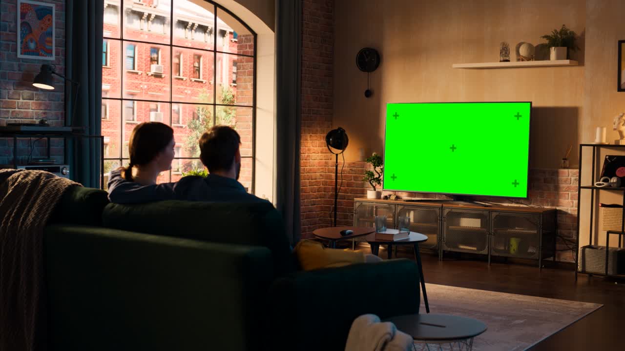 젊은 부부는 소파에 앉아서 집에서 시간을 보내고, 그들의 세련된 로프트 아파트에서 녹색 화면 모의 디스플레이를 가진 tv를보고 있습니다. 남자와 여자는 영화 또는 쇼를 스트리밍하고 팝콘을 먹습니다.