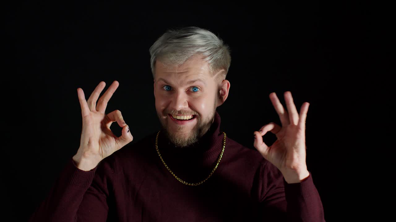 금발과 수염을 가진 젊은 남자가 미소 짓고 손으로 은 표시를 합니다.