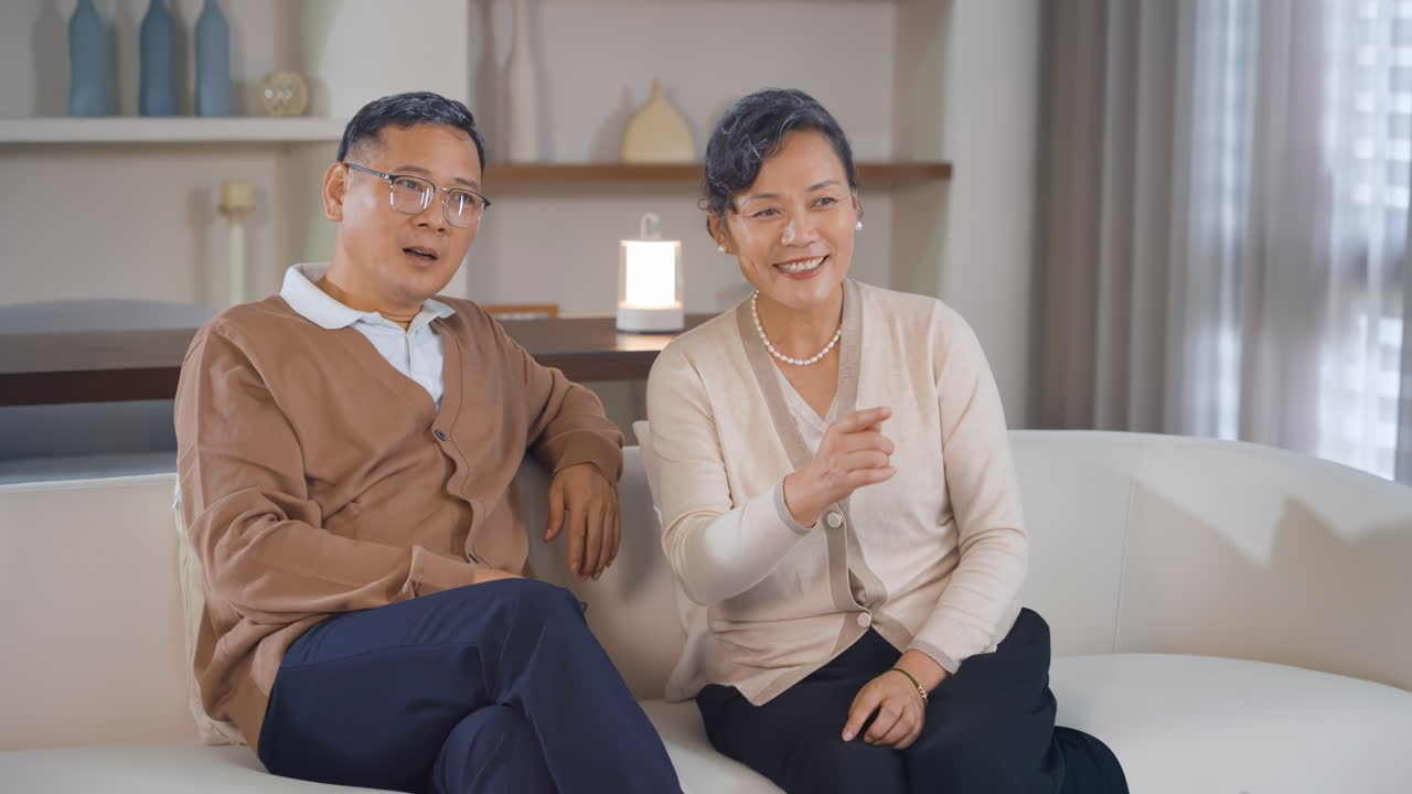 คู่สามีภรรยาสูงอายุที่มีความสุขกำลังหัวเราะและดูทีวีด้วยกันในวัยเกษียณ พูดคุยกันพร้อมถือรีโมทคอนโทรล แนวคิดวิถีชีวิตครอบครัวและการพักผ่อนของชายหญิงสูงอายุที่มีรอยยิ้มและโทรทัศน์