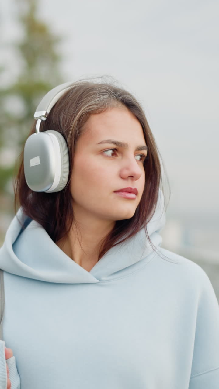 Close-up view of student walking calmly while listening to music with headphones in hoodie, blurred background features street lights and withered trees, peaceful urban scene