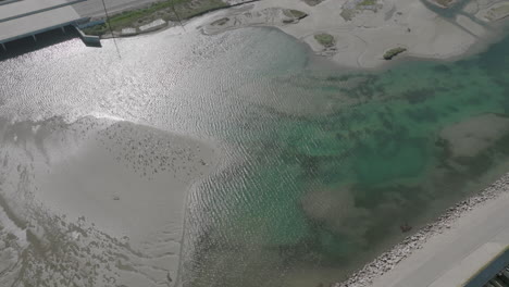 Sobrevuelo-Aéreo-De-Un-Río-Junto-Al-Océano,-Con-Un-Puente-Y-Coches-Conduciendo-Al-Fondo