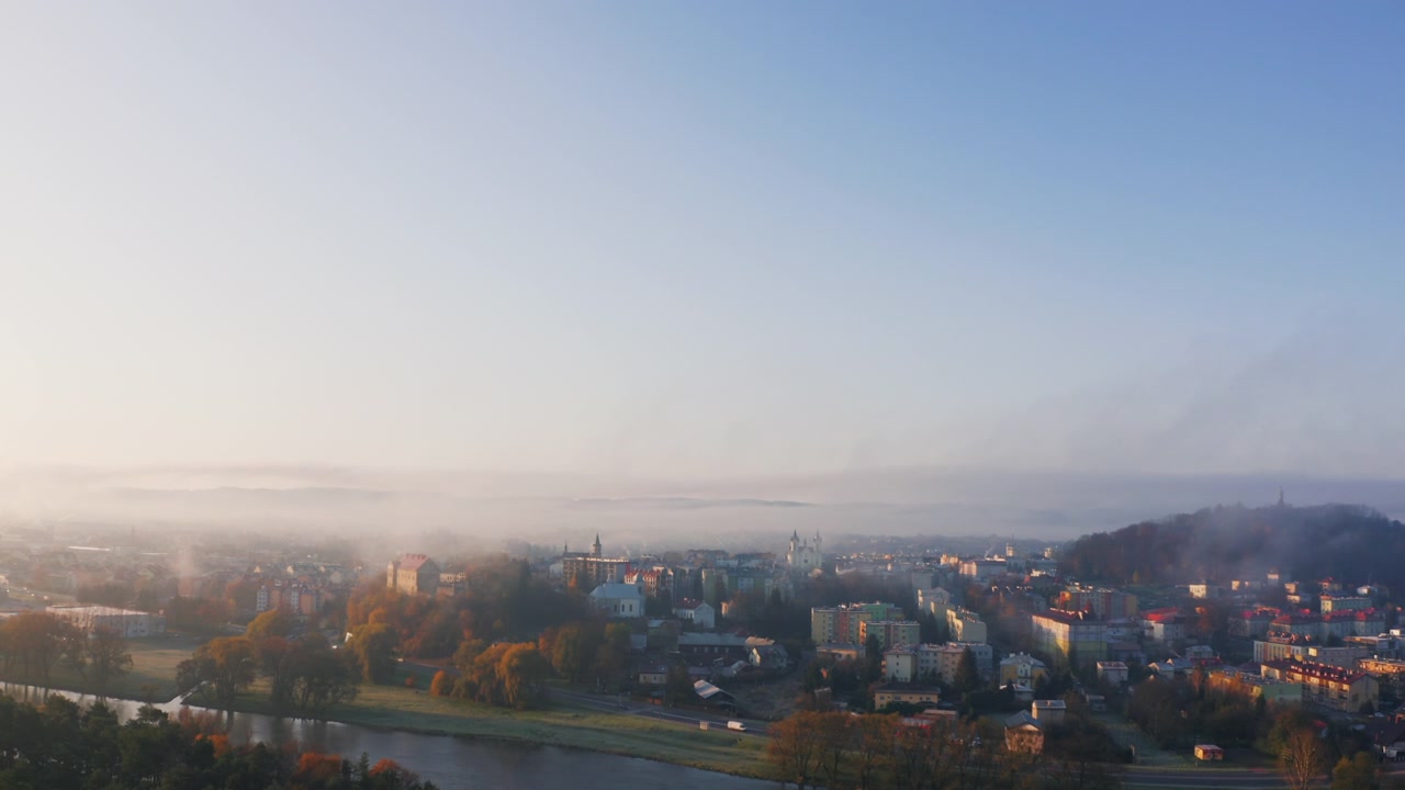 Syksyinen drone-kuva sumun peittämästä kaupungista ja joesta auringonnousun aikaan, kylpien pehmeässä aamun hehkussa. Valo valaisee sumun luoden lämpimän, eloisan, ainutlaatuisen maiseman. Sininen taivas lisää värikontrastia. Sanok.