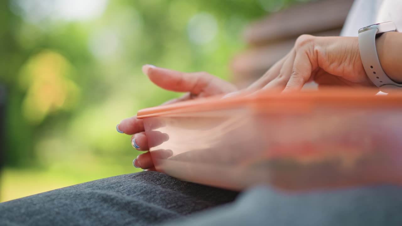 Una mujer blanca aficionada al fitness abre una fiambrera naranja en un banco para coger lechuga y un sándwich después de entrenar, primer plano de las manos y el reloj con un fondo de vegetación iluminada por el sol, momento espontáneo de descanso activo