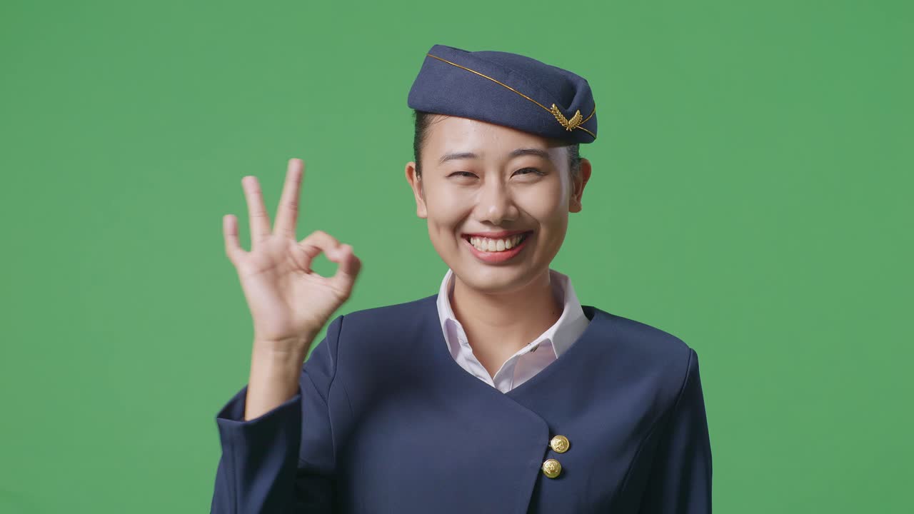 녹색 스크린 배경 스튜디오에 서있는 동안 미소 짓고 카메라에 은 몸짓을 보여주는 아시아 여성 항공 승무원