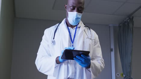 médico afroamericano que usa una máscara facial usando una tableta digital en la habitación del paciente del hospital