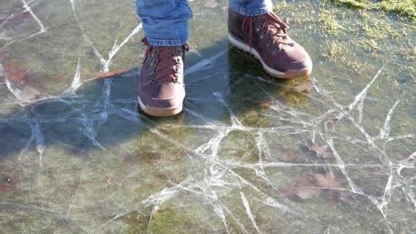 close up of man's feet wearing brown boots. standing on a frozen pool over grass in a park on a sunny winter day. feet slide sideways until reaching the grass and then he walks away.