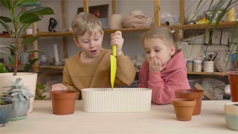 little blonde girl and blond kid preparing the soil in a pot sitting at a table where is plants in a craft workshop