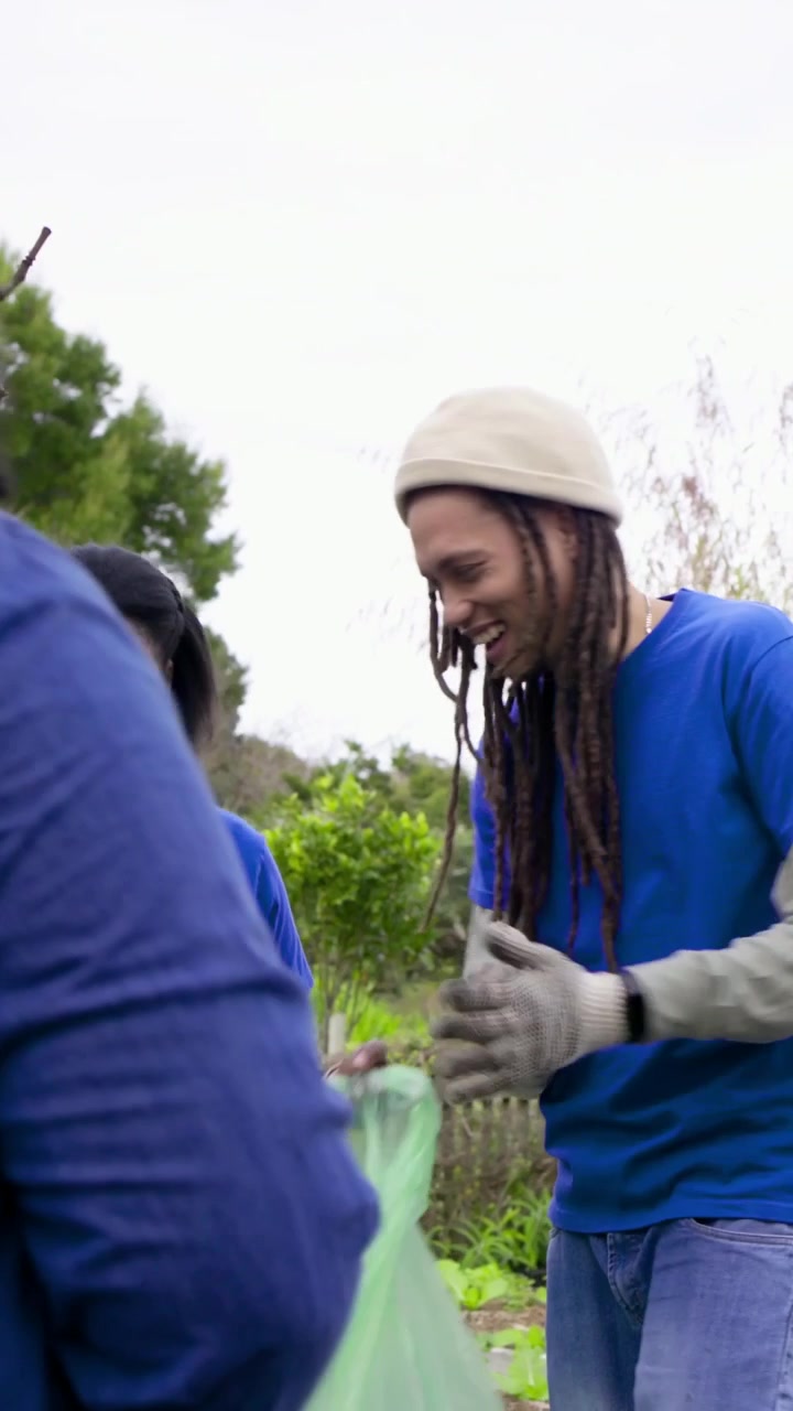 Video verticale: Apertura di un sacco verde, una donna e una squadra con magliette blu che rimuovono detriti in un orto comunitario.