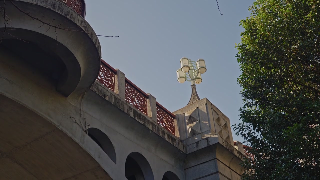 Ripresa statica dal basso di un parapetto di un ponte storico in pietra, ringhiera a traliccio rossa che curva a sinistra, elegante lampione a più globi centrato contro un cielo azzurro limpido, fogliame verde degli alberi che incornicia il bordo destro.