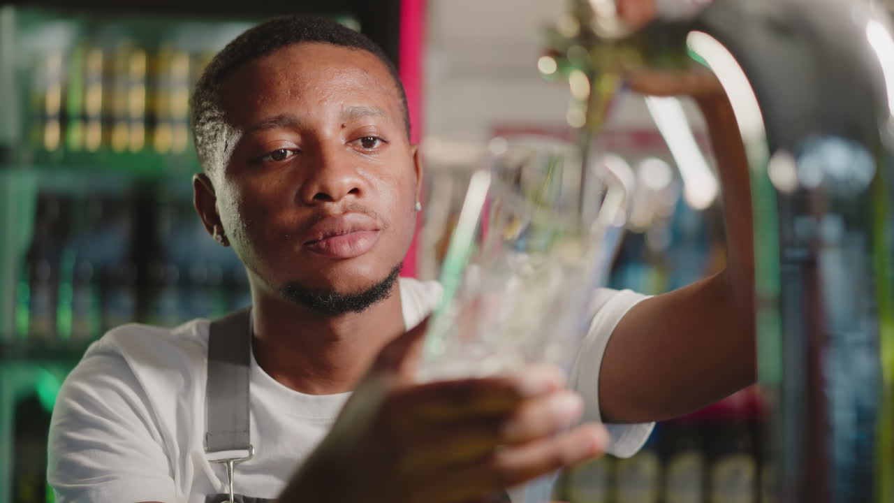 Barman fills glass with beer in pub closeup. Serious African American bartender pours ale into mug working at counter. Black guy serves drinks