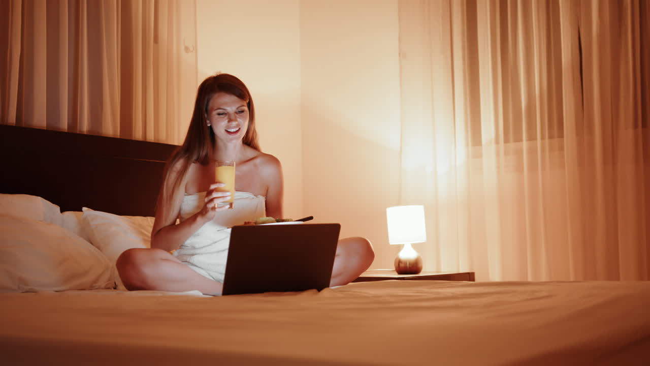 밤에 침대에서 주스를 마시며 노트북을 사용하는 여자