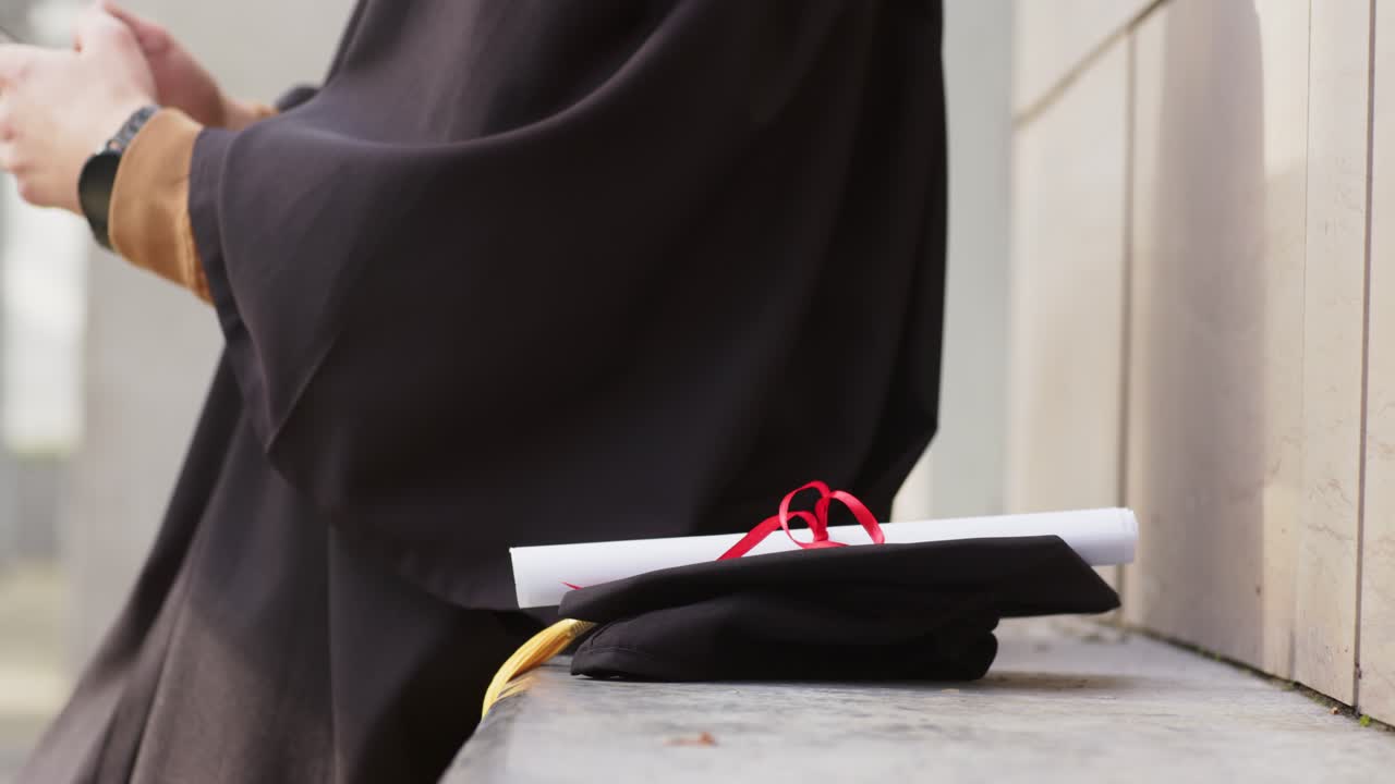Dopo aver posizionato il tocco e il diploma sulla panchina, laureato in toga che prende lo smartphone, controllando le notifiche