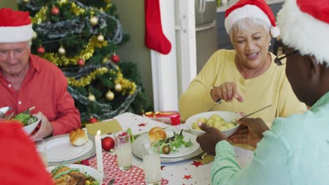 group of happy diverse senior friends in santa hats passing food at christmas dinner table at home