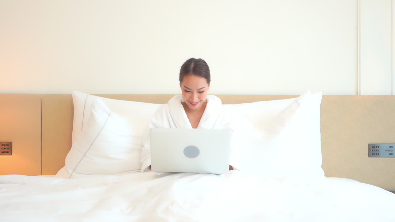 집에서 일하고, 밝은 아침에 노트북 컴퓨터에 목욕 가운을 입고 침대에 누워 있는 예쁜 아시아 여성, 풀 프레임
