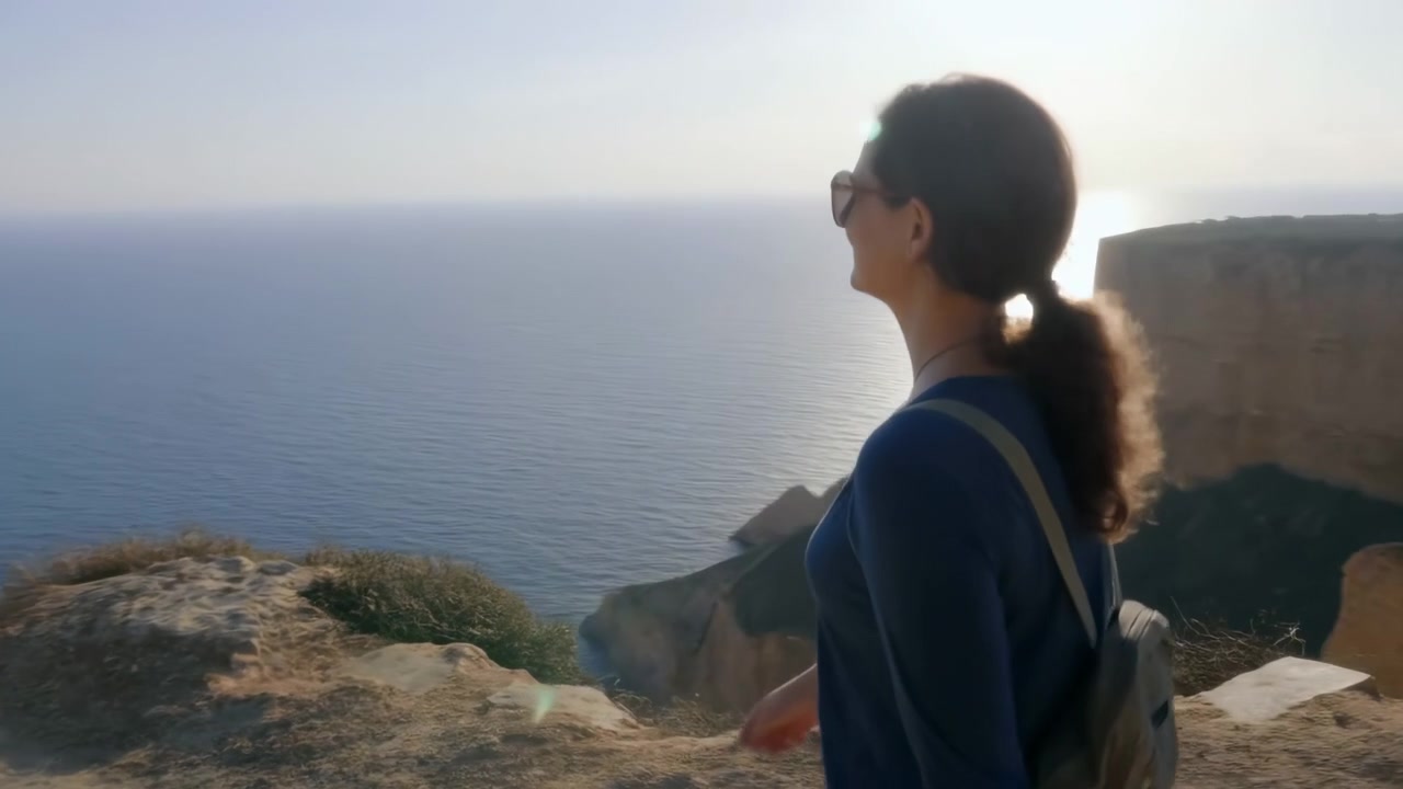 Viste laterali e posteriori seguono una donna che passeggia oltre un terreno roccioso e arido e ripide scogliere marine, con acqua blu calma, orizzonte lontano, luce solare calda e un leggero bagliore dell'obiettivo dietro di lei.