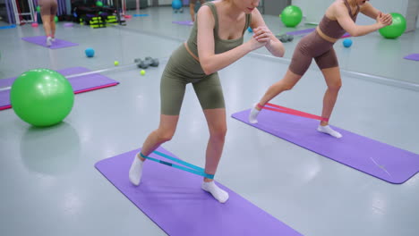 mujeres que participan en un entrenamiento de fitness usando bandas de resistencia en esteras de yoga púrpura en un gimnasio, se centran en ejercicios de fortalecimiento y estabilidad de la parte inferior del cuerpo, con equipos de gimnasio visibles en el fondo
