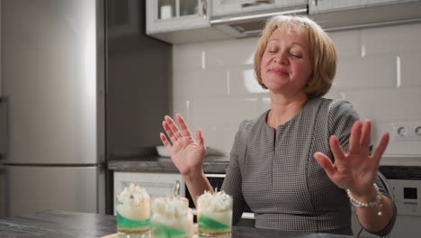 Female baker seated in kitchen in front of baked desserts sways rhythmically with eyes closed while demonstrating expressive hand dance movements in joyful moment celebrating culinary success
