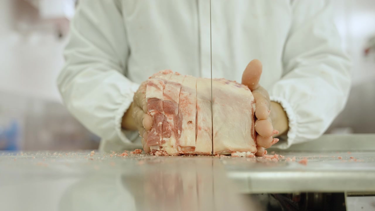 Gloved butcher hands push a large lamb rib section through a vertical bandsaw, cutting even portions on a stainless table.