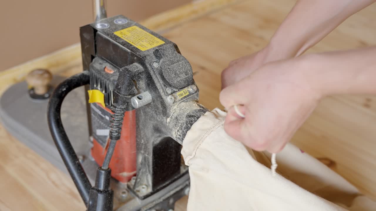 Closeup of person attaching dust collection bag onto sanding machine on wood floor edge finishing trim beside wall