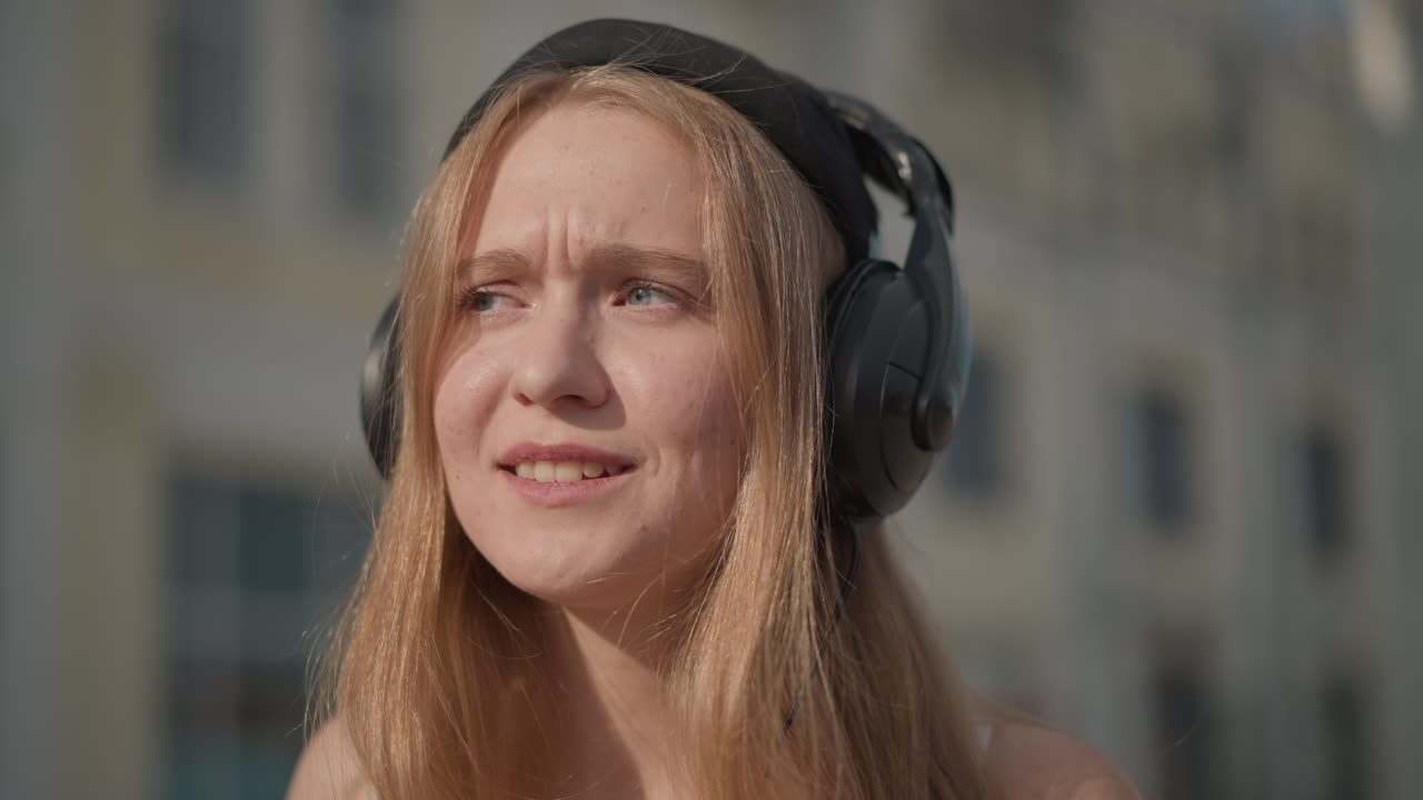 Donna con cuffie in una strada illuminata dal sole, sorridente, che sistema un berretto nero, capelli dorati illuminati dal sole, ritratto urbano spontaneo, leggero movimento a ritmo di musica, primo piano che cattura l'espressione gioiosa e naturale