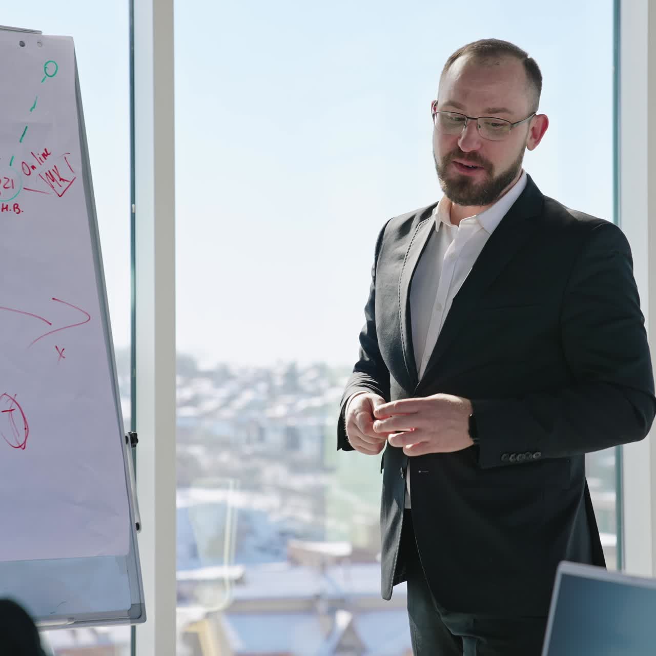 Male speaker explaining the chart beside him and gesturing actively in front of the team. Big windows and cityscape at the backdrop