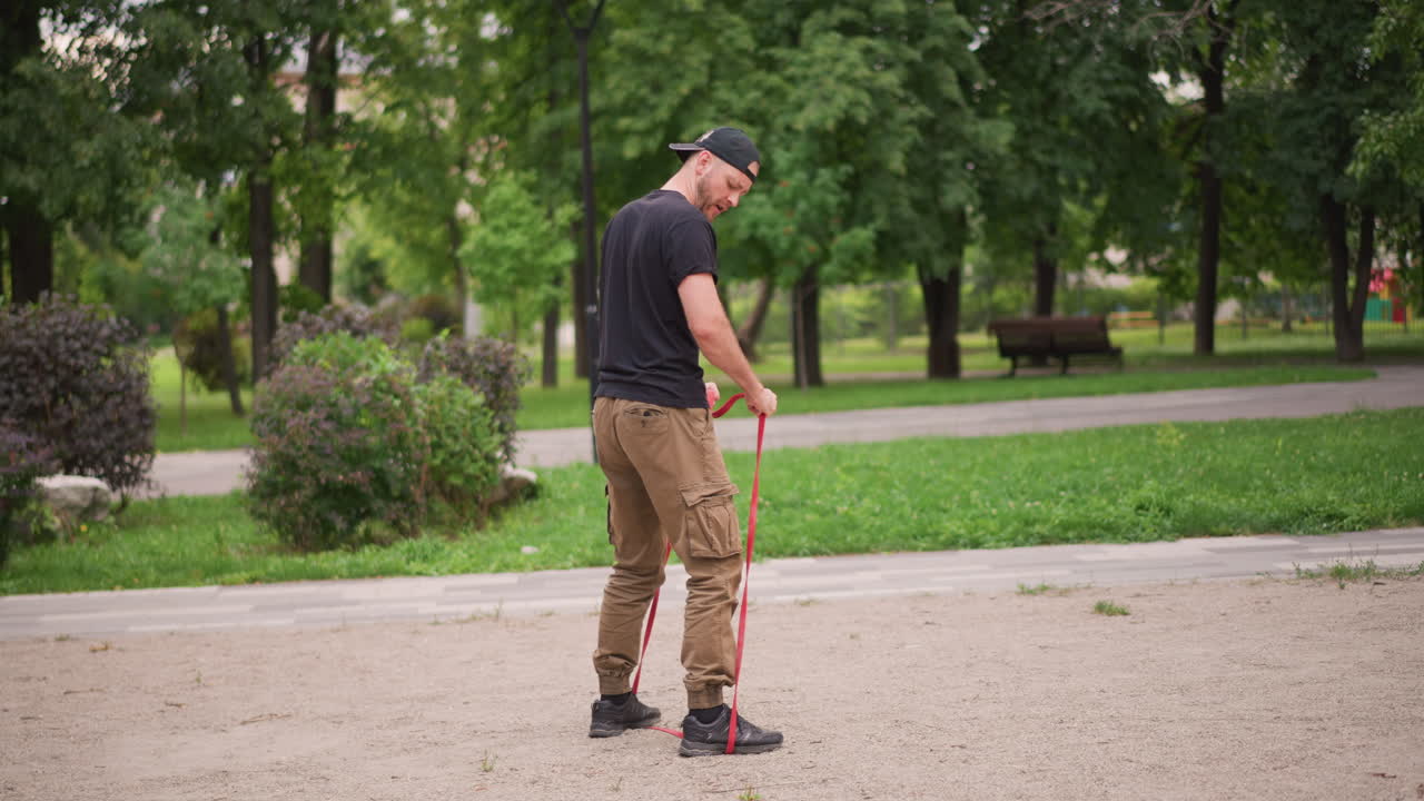 Uomo bianco con fascia di resistenza fissata al piede che esegue esercizi di stepping, pantaloni cargo e berretto, focus sulla spinta della caviglia e allenamento della forza esplosiva in un ambiente esterno verdeggiante
