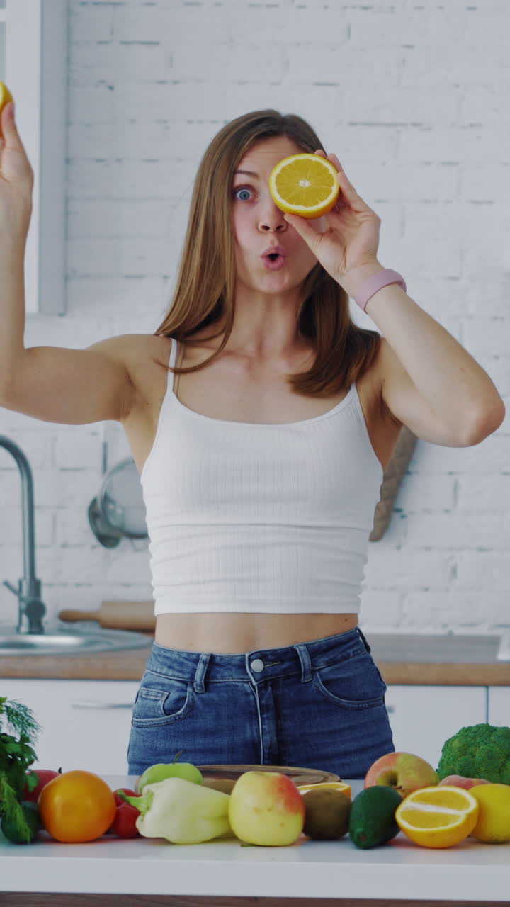 Young woman with oranges in the kitchen and many fresh fruit and vegetables on the table. Beautiful female holding two halves of orange closing her eyes and looking at camera merrily. Vertical video