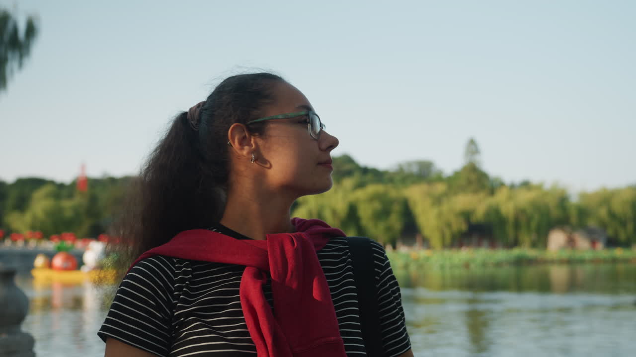 Woman with glasses by lake during golden hour, closeup portrait, red scarf draped over shoulders, polka dot top, soft sunlight, calm water background, distant boats and green trees, peaceful