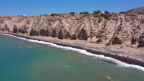 Aerial-flying-across-Greek-Island,-rocky-cliff-mountains-and-blue-water-waves-in-the-Mediterranean-Sea-on-white-beach-in-Santorini,-Greece