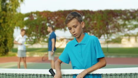 Porträt-Eines-Müden-Teenagers-Mit-Schläger,-Der-Sich-Auf-Dem-Netz-Lehnt-Und-Auf-Einem-Tennisplatz-In-Die-Kamera-Lächelt