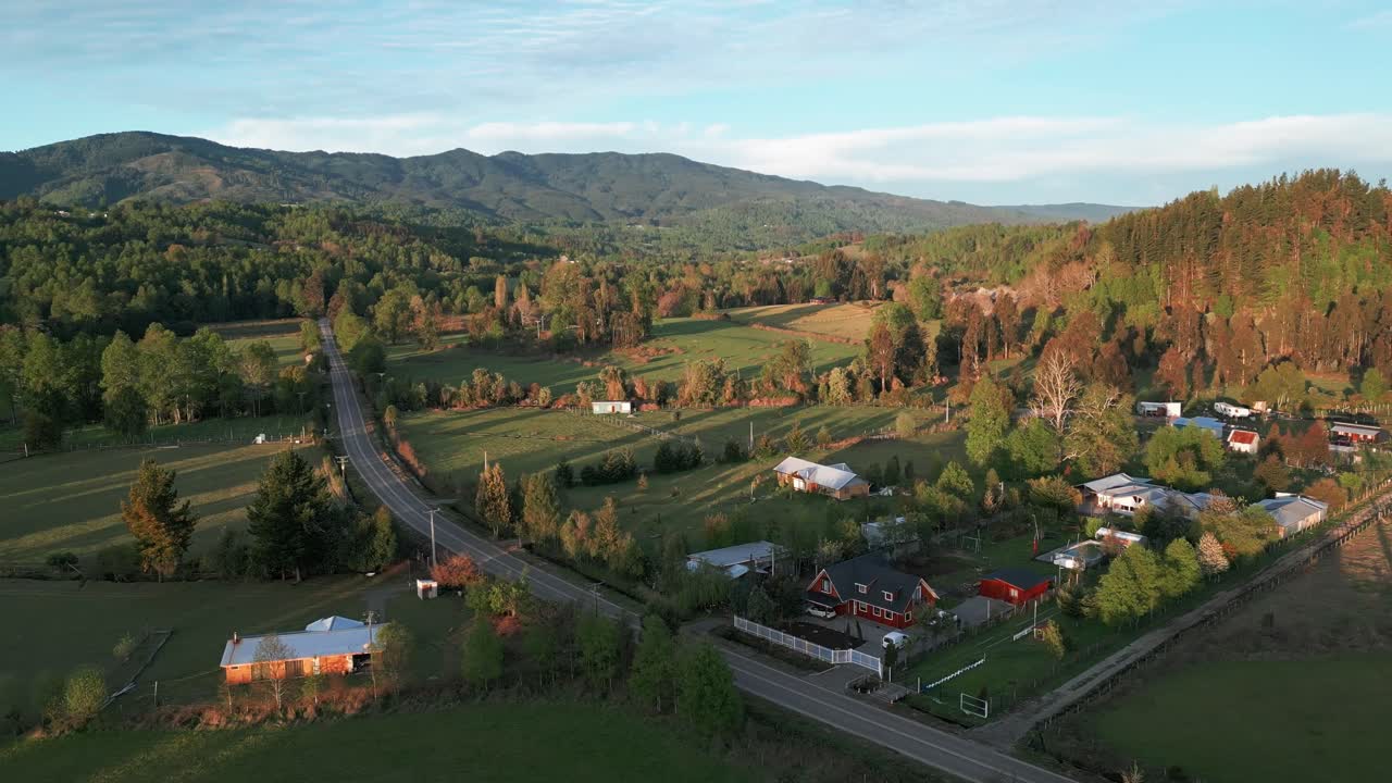 Vista aerea da drone orbitante di un villaggio rurale con case, strada tortuosa e fogliame autunnale immerso tra campi, foreste e paesaggi montani ondulati