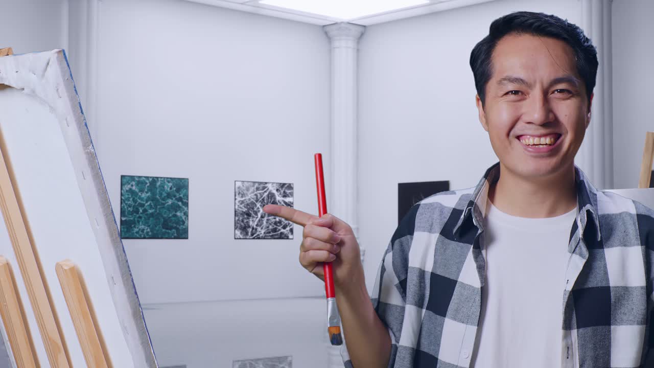 갤러리에서 오일 페인트와 브러쉬로 그림을 그리는 동안 캔버스를 가리키는 아시아 남성 예술가의 클로즈업