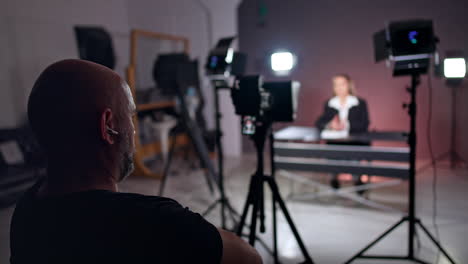 bald neared man sits leaned back in the chair. rear view of a cameraman sitting in studio behind the camera. caucasian woman sits at blurred backdrop.